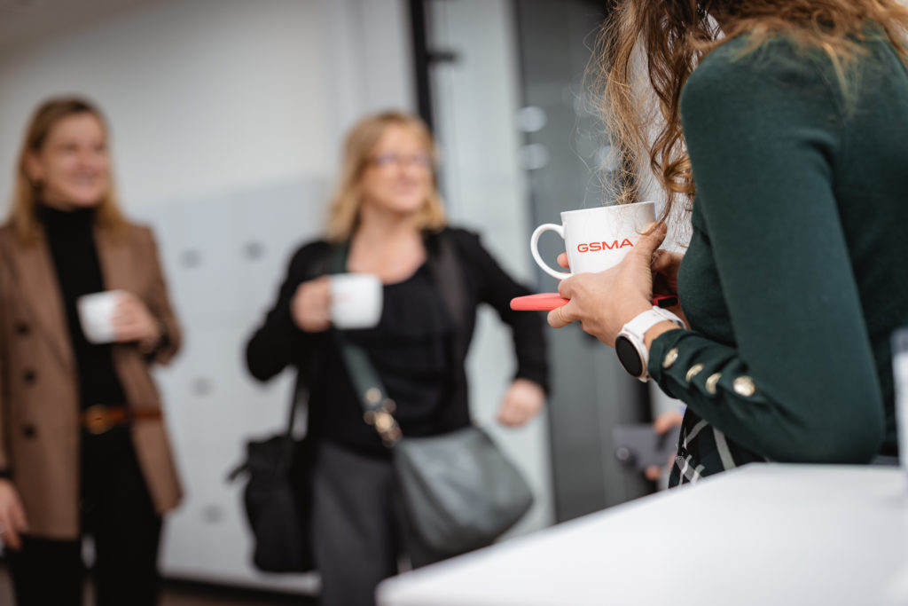 Three women are casually conversing in an office setting, holding white mugs with the "GSMA" logo. The focus is on the woman in the foreground, who wears a green sweater and has a smartwatch on her wrist. The background is slightly blurred, emphasizing a relaxed atmosphere.