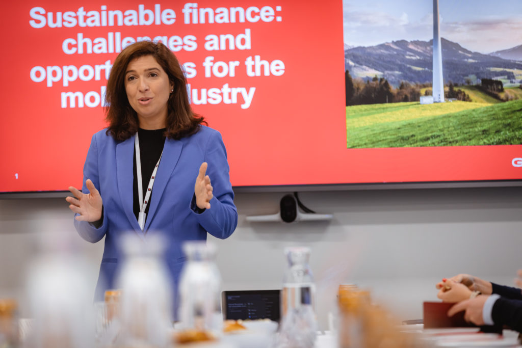 A woman in a blue blazer speaks animatedly in front of a presentation slide titled "Sustainable finance: challenges and opportunities for the mobile industry." The slide features a landscape with mountains and a wind turbine. People seated in the foreground are partially visible.