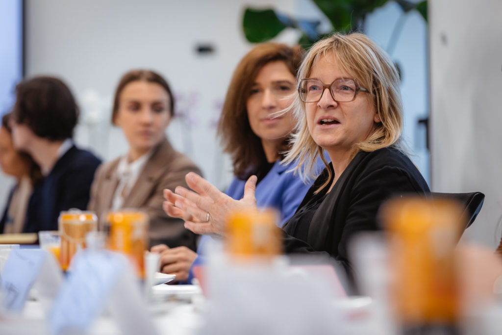 A woman with glasses speaks animatedly at a conference table. She is surrounded by others, who are listening. There are nameplates and drinks on the table, and a plant in the background. The scene suggests a professional discussion or meeting.