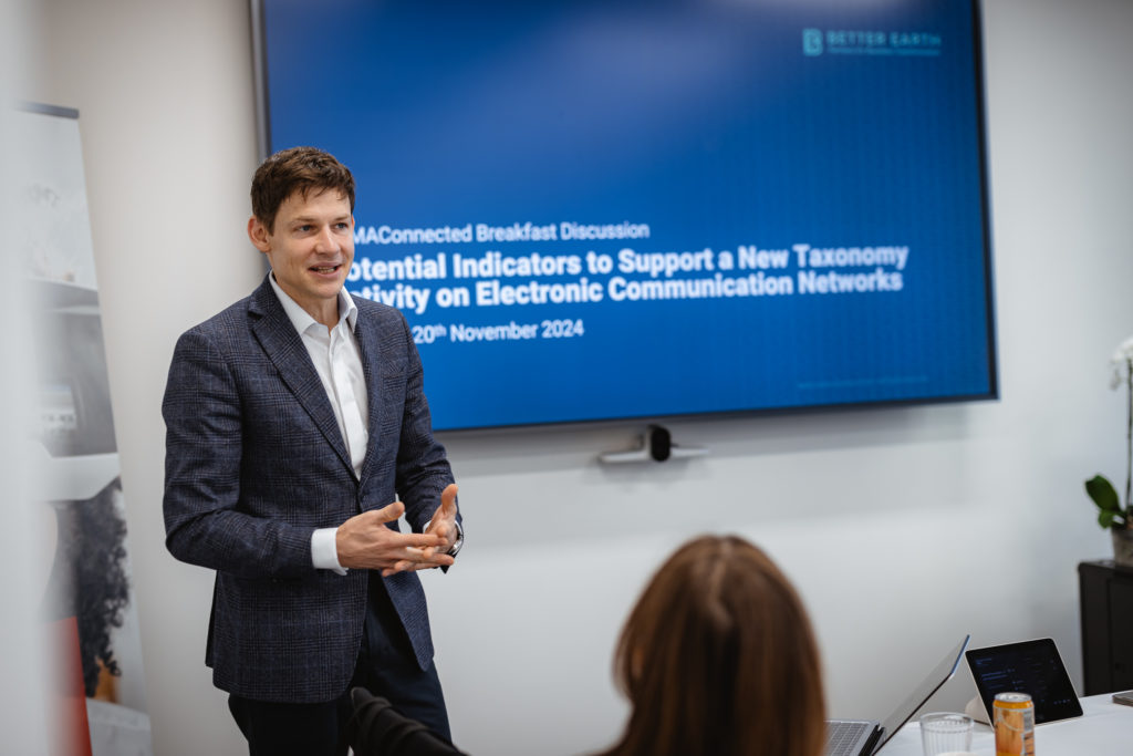 A man in a suit gestures while speaking in front of a presentation screen. The screen displays the title "Potential Indicators to Support a New Taxonomy: Connectivity on Electronic Communication Networks" with the date "20th November 2024." A woman is partially visible, listening.