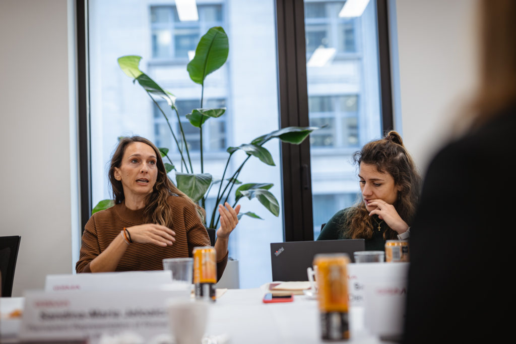 Two women are sitting at a conference table in an office setting. One is speaking with gestures, while the other types on a laptop. Drink cans and papers are on the table. Large windows in the background show a cityscape. A potted plant is visible near the windows.