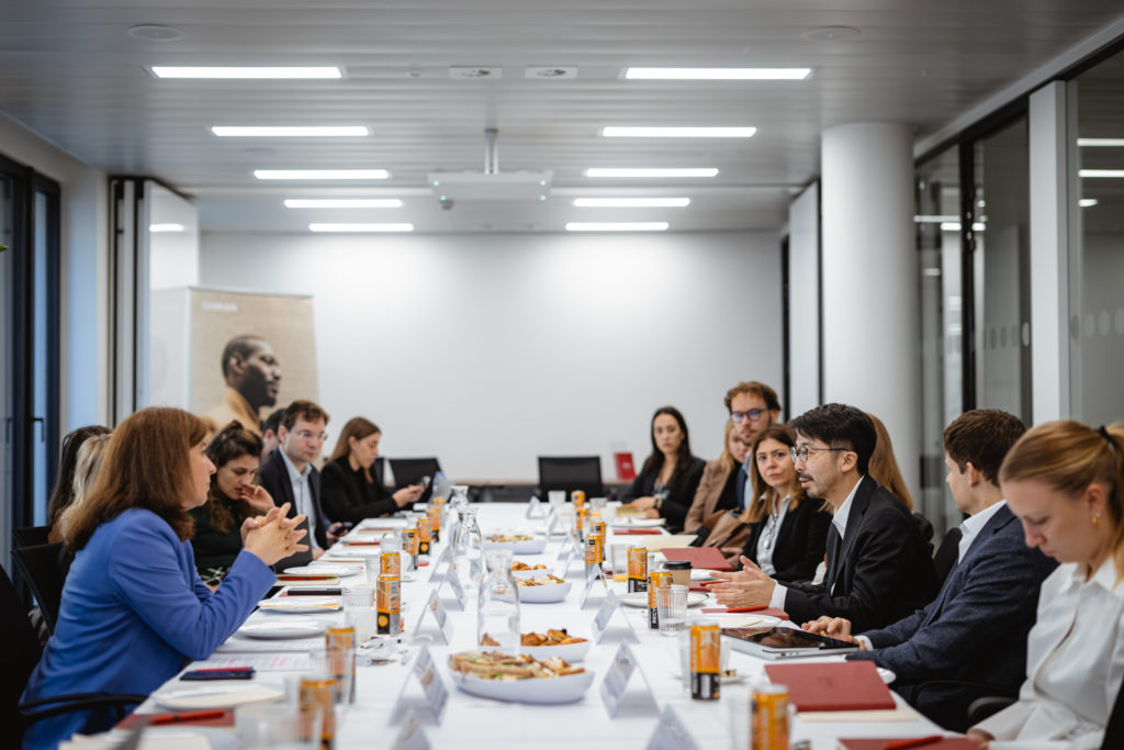 A diverse group of professionals in business attire sit around a long conference table in a modern office setting. Papers, notebooks, and drinks are visible on the table. A large poster and glass walls are in the background, creating an open atmosphere.