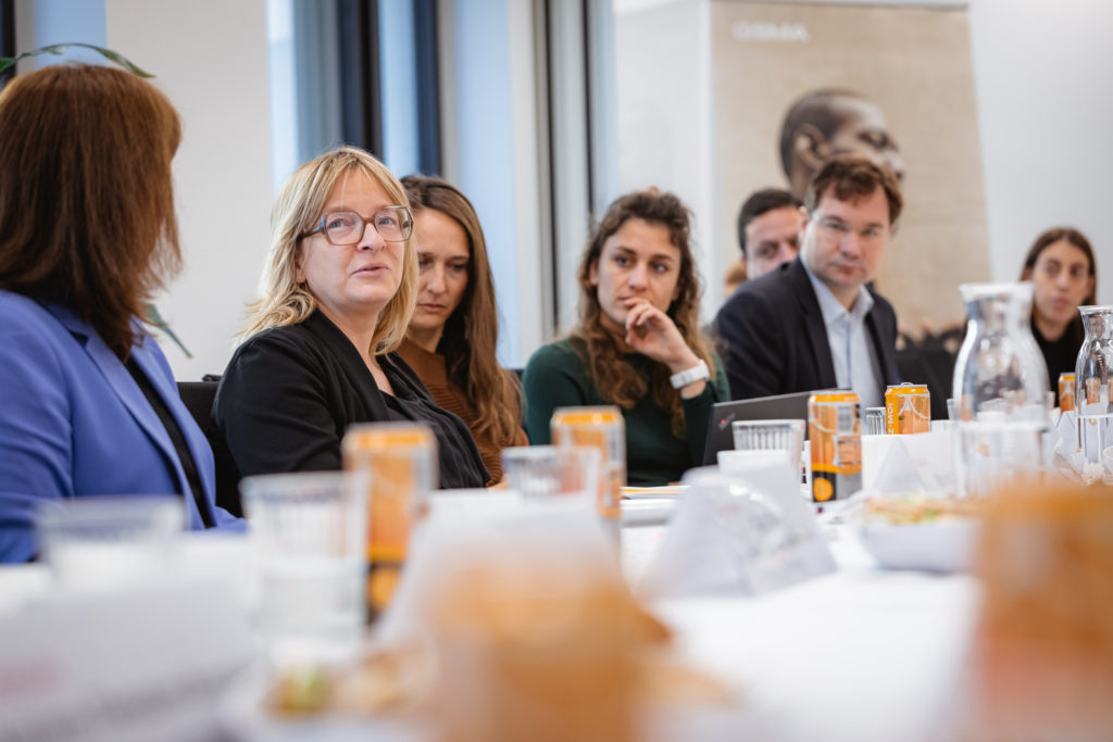 A group of people sit around a table in a meeting room. Drinks, papers, and nameplates are on the table. Various individuals are engaged in conversation, with some listening attentively. A poster is visible in the background. Large windows allow natural light into the space.