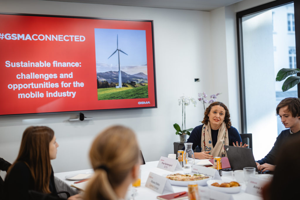 A group of people are seated around a conference table in a meeting room. A large monitor displays a slide titled "#GSMA CONNECTED: Sustainable finance: challenges and opportunities for the mobile industry," featuring a wind turbine. The participants are engaged in discussion.