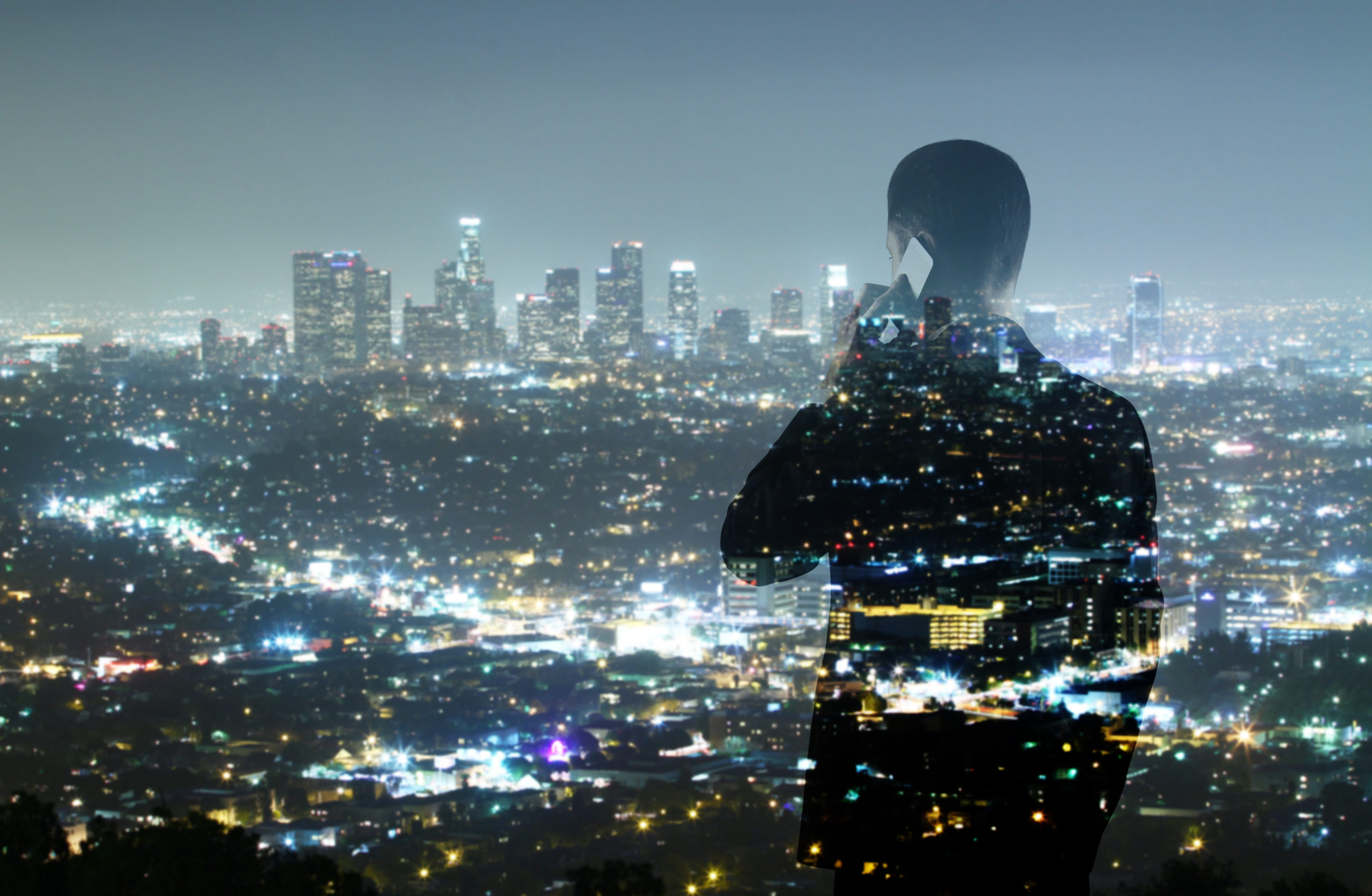 Silhouette of a person on a phone, overlayed with a cityscape at night. The skyline features illuminated skyscrapers and dense streetlights, creating a dynamic interplay of lights and urban structures against a clear night sky.