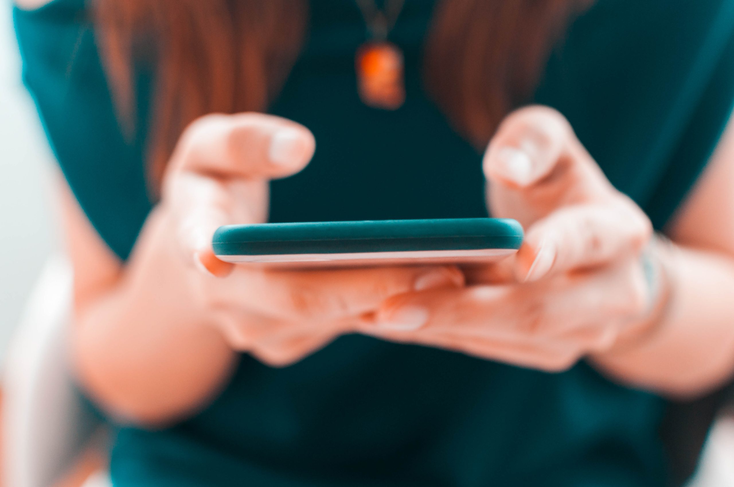 A person holding a smartphone with both hands, focusing on the screen. The individual is wearing a dark green top, and a necklace with an orange pendant is visible. The image is slightly blurred, emphasizing the action of using the phone rather than the person's features.