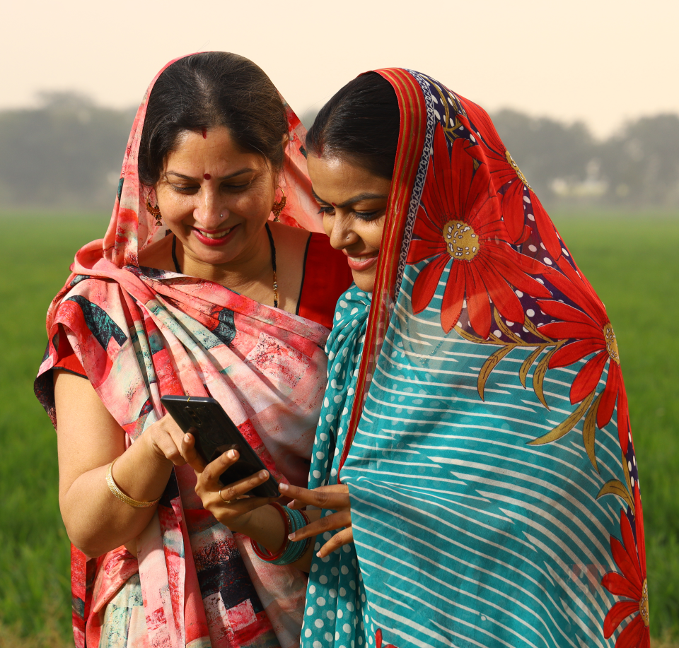 Two women in brightly colored sarees, standing together in a field, smile as they look at a smartphone. The woman on the left wears a pink and white saree, while the woman on the right is in a blue saree with red flower patterns. Trees are visible in the background.