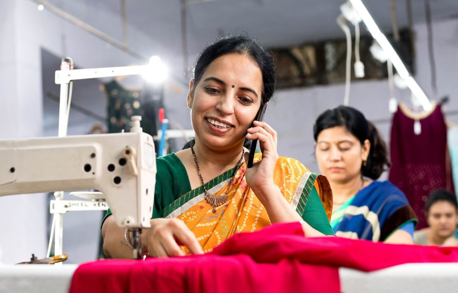 A woman in a colorful sari smiles while talking on a phone and sewing with a sewing machine in a workshop. Another woman in the background, also dressed in a sari, focuses on sewing. Fabrics and clothing hang in the background. Bright lighting illuminates the workspace.