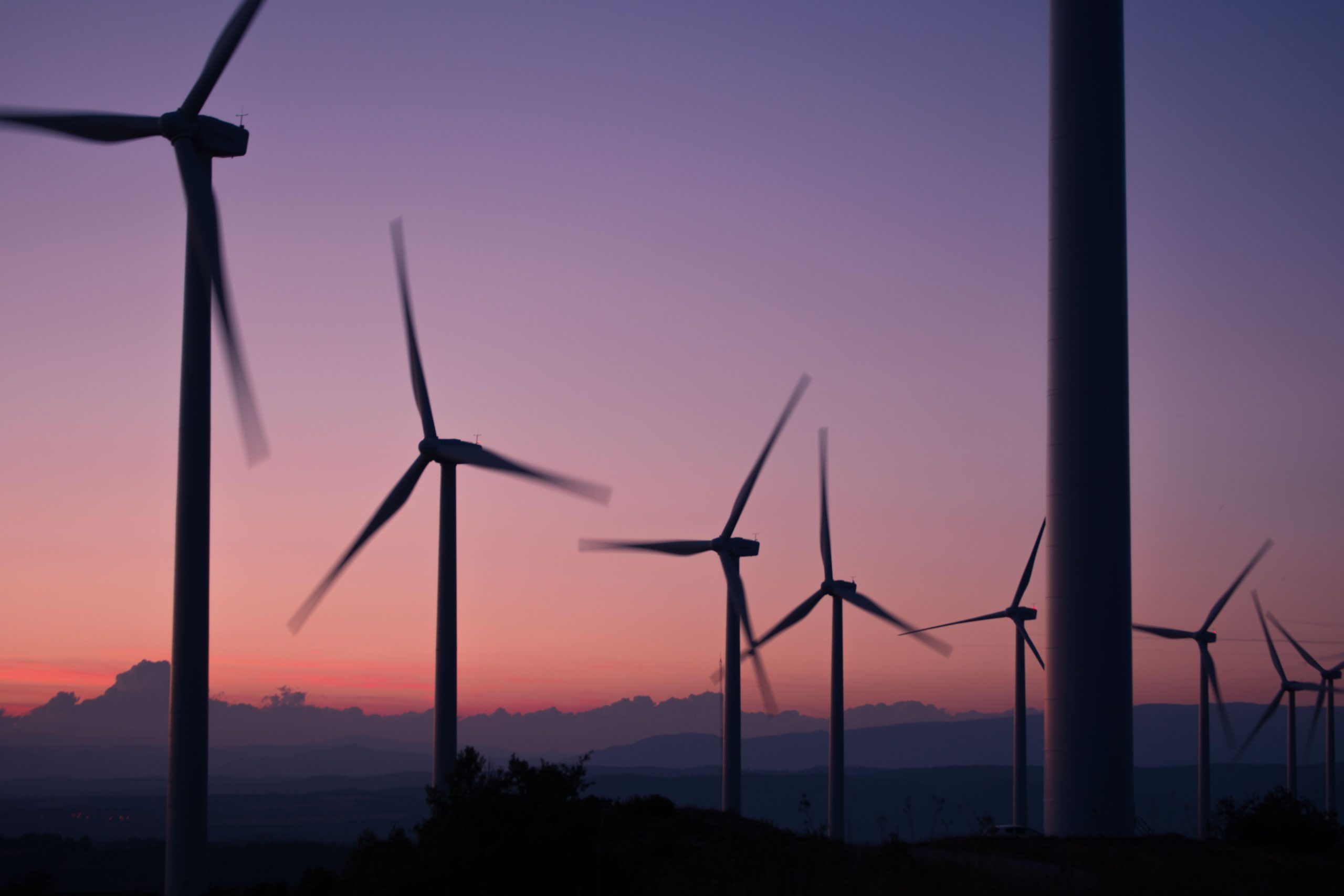 A row of wind turbines is silhouetted against a sunset sky, with mountains in the background. The blades are blurred, indicating motion. The sky transitions from pink to purple as the sun sets.