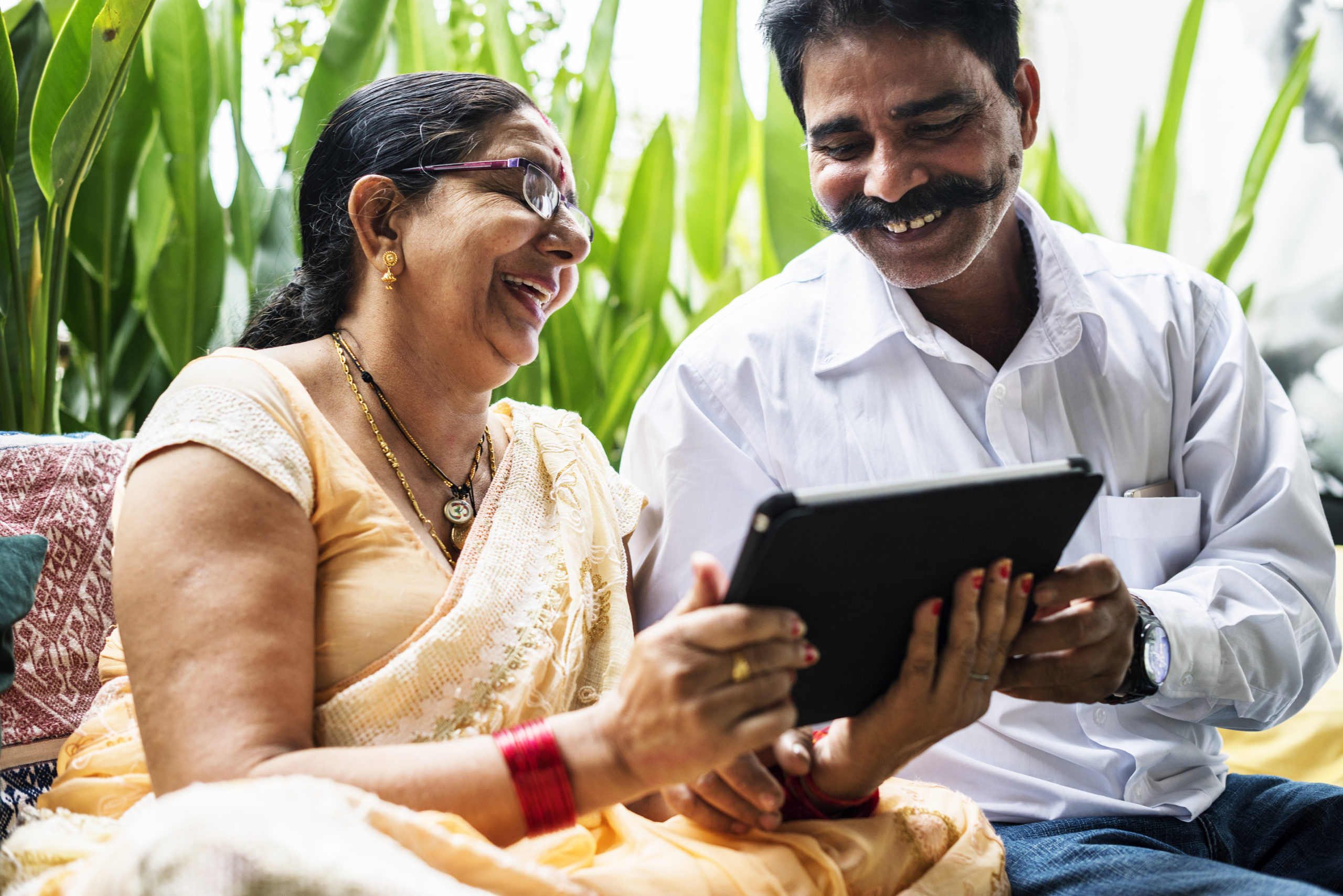 A smiling woman in a saree and a man with a mustache, both wearing glasses, are sitting together outdoors. They are looking at a tablet screen and laughing. Plant leaves are visible in the background.