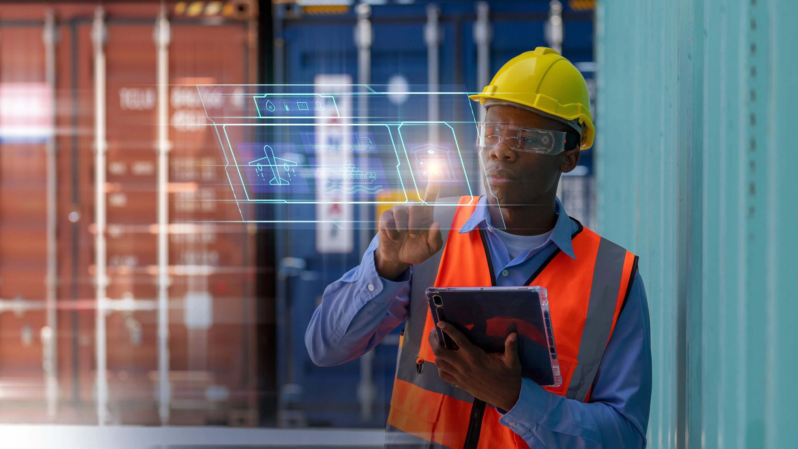 A person wearing a yellow hard hat, safety goggles, and an orange safety vest is interacting with a transparent digital interface. They are holding a tablet in one hand and standing in front of shipping containers. The digital interface displays cargo information.