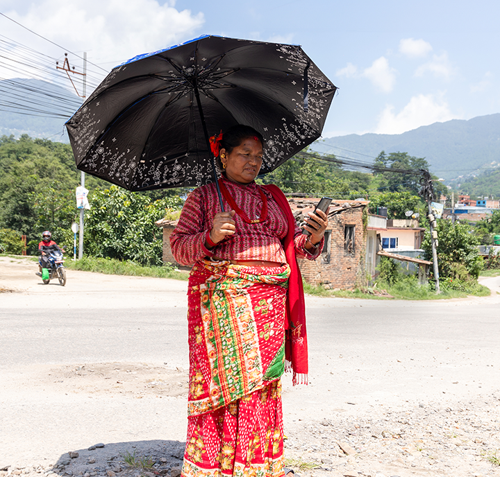 A connected woman wearing a red traditional outfit and holding a black umbrella stands at a roadside, looking at her phone. The background includes mountains, buildings, and a motorcycle in motion.