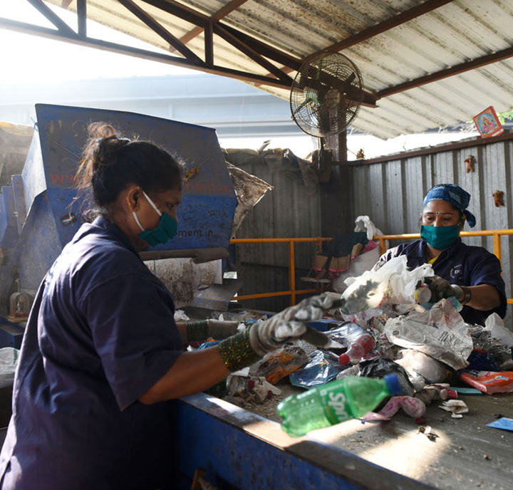 Two workers wearing masks and gloves sort through waste at a recycling facility. They stand at a conveyor belt, separating items while a large machine and an overhead fan are visible in the background. The scene, reminiscent of innovations supported by GSMA Mobile for Development, is lit by natural light.