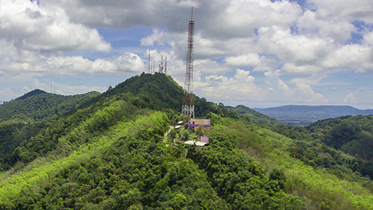 Aerial view of a lush green hilltop with multiple communication towers and a small cluster of buildings under a partly cloudy sky.