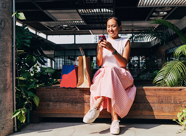 A woman sitting on a bench, using her smartphone with shopping bags beside her, smiling, outdoors.