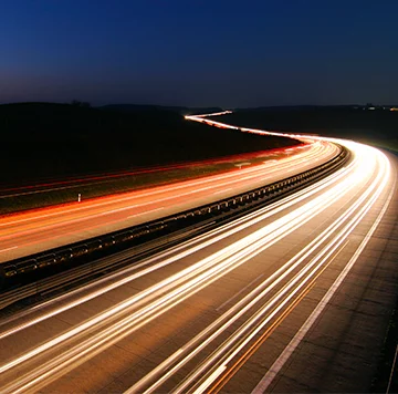 A long-exposure photo of a highway at night. Headlights and taillights of fast-moving vehicles create continuous streams of white and red light, giving the impression of speed and movement against a dark, clear sky. The roadway is mostly empty with minimal surrounding details visible.