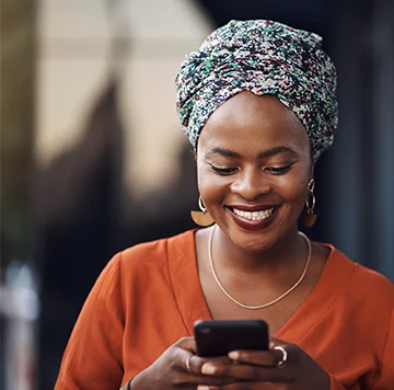 A woman with a beaming smile looks down at her smartphone. She wears an orange top, large hoop earrings, and a colorful headwrap. The background is softly blurred, drawing focus to her joyful expression and poised appearance as she engages with the device in her hands.