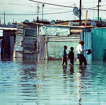 Three people, an adult and two children, wade through knee-deep floodwater in a makeshift settlement. The area is filled with temporary structures made from corrugated metal and wood. Satellite dishes and power lines are visible above. The sky is overcast.