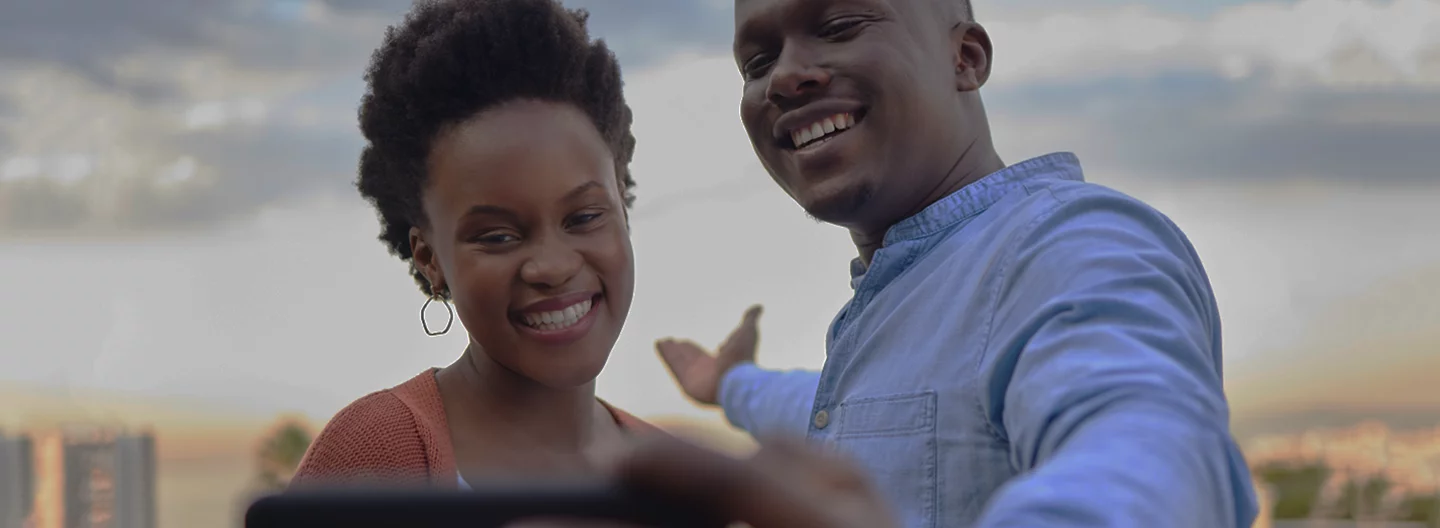 A smiling couple takes a selfie outdoors. The woman, wearing a reddish cardigan and hoop earrings, stands beside the man, who is in a blue shirt. Both appear happy, with the man gesturing with his arm. The background shows a cityscape under a cloudy sky during sunset.